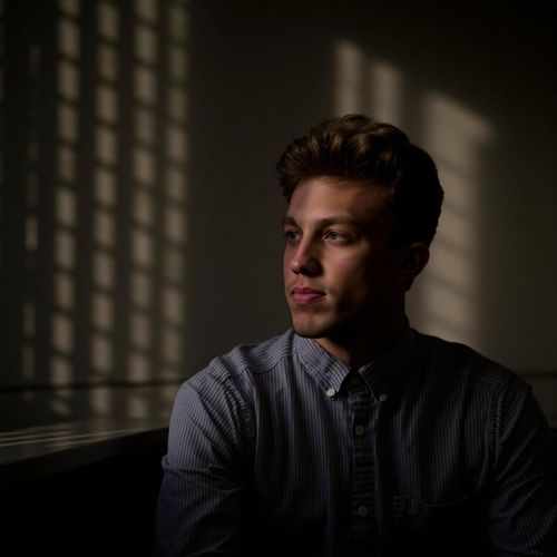 a photo of me MAN in a dimly lit office, half body shot, shadows from window blinds striping the scene. MAN looking thoughtful.