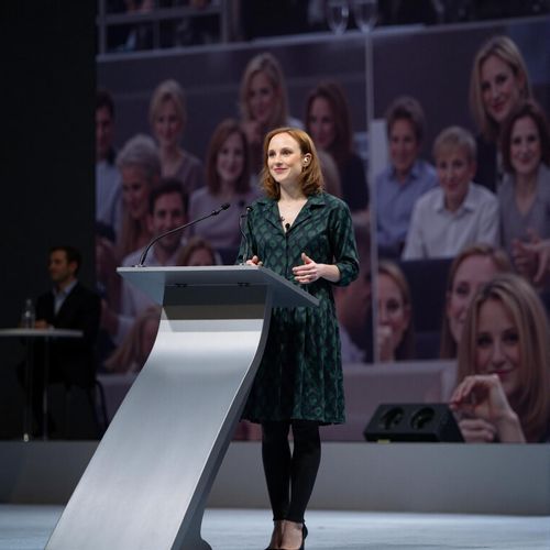 Showcase image for On The Main Stage - Prompt: a photo of me WOMAN as a keynote speaker, medium shot, standing confidently at a sleek podium on a large stage, blurred audience in far distance. WOMAN looking authoritative.